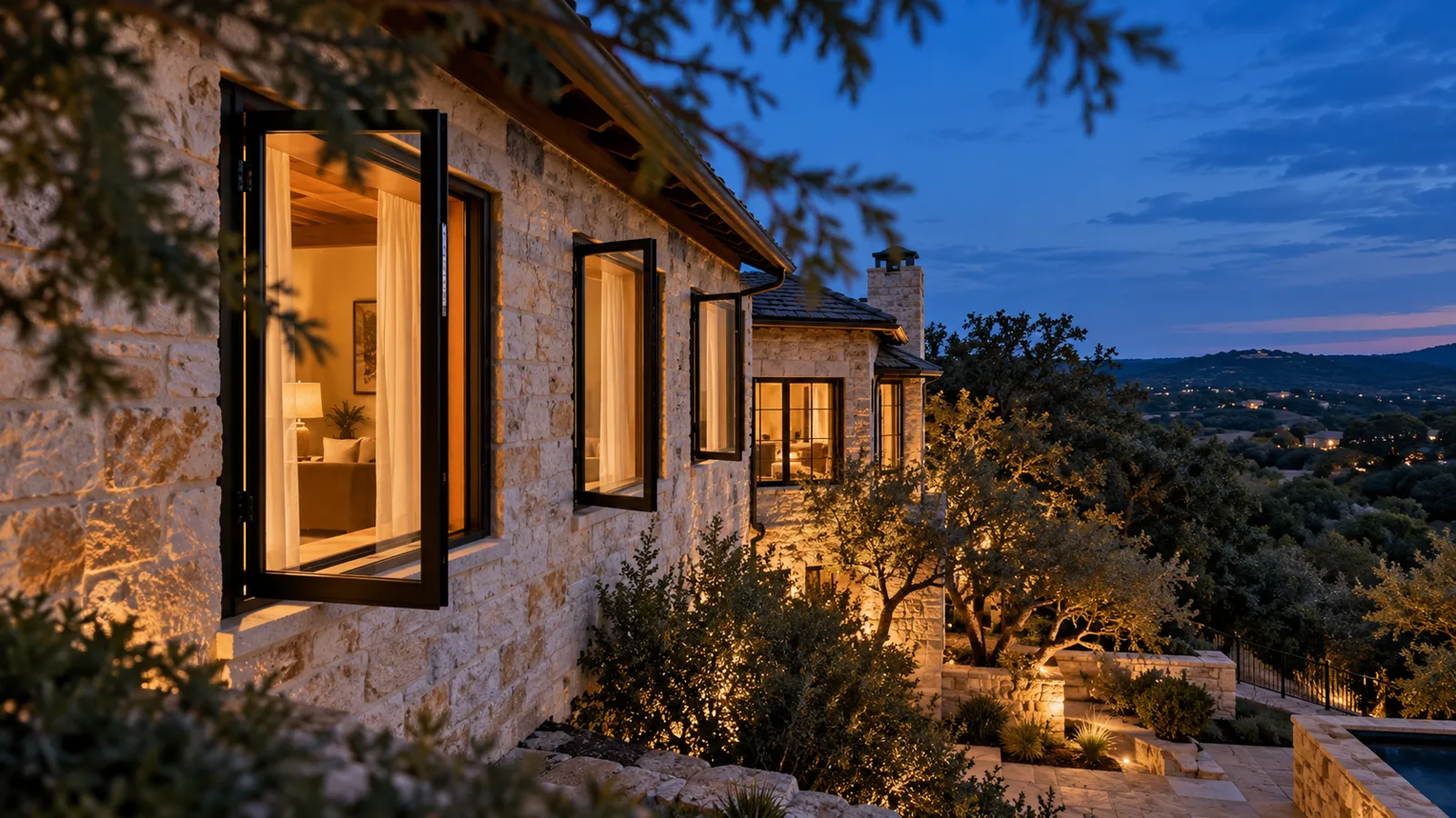Black crank-out casement windows open on a limestone Hill Country home near Austin at dusk with warm lights inside