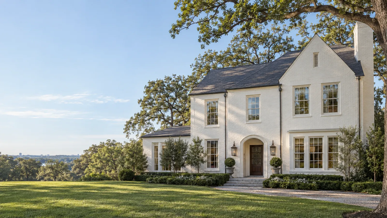 A white-painted brick house with tall divided-lite double-hung windows under mature oak trees